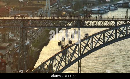 Bei Sonnenuntergang gleiten Ragelo-Boote entlang des goldenen Flusses Douro unter der Dom Luis I Brücke, mit Fußgängern auf der Brücke und Reflexionen an der Uferpromenade in Porto und Gaia. Drohnenaufnahme aus der Luft Stockfoto