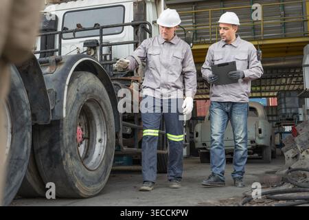 Kaukasische und mittelgroße männliche Techniker aus dem Nahen Osten gehen durch einen asiatischen Containerhof, während sie ein Klemmbrett halten, das für Routineinspektionen steht Stockfoto