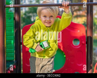 Der blonde Junge in grünem Hoodie lächelt, während er den Spielzeugwagen an sonnigen Tagen auf einem farbenfrohen Spielplatz hält Stockfoto