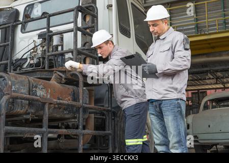 Kaukasische und mittelgroße männliche Techniker aus dem Nahen Osten gehen durch einen asiatischen Containerhof, während sie ein Klemmbrett halten, das für Routineinspektionen steht Stockfoto