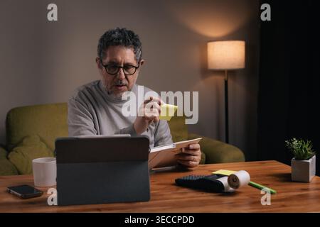 Mann mittleren Alters, der persönliche Aufgaben mithilfe von Haftnotizen organisiert, während er abends zu Hause am Tablet arbeitet. Produktivitäts-, Zeitmanagement-, Stockfoto