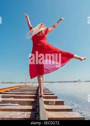 Eine junge Frau in rotem Kleid läuft barfuß auf Eisenbahngleisen über den Salzsee Bursol unter klarem blauem Himmel Stockfoto