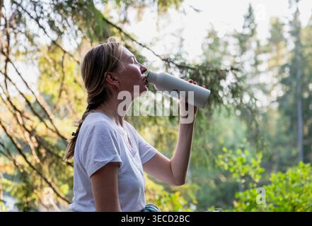 Junge Frau trinkt Wasser aus wiederverwendbarer Flasche im Wald während der Sommeraktivität im Freien Stockfoto