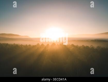 Die Luftaufnahme fängt den Sonnenaufgang über einem nebeligen Waldtal ein und entfernte Hügel erzeugen dramatische Lichtstrahlen Stockfoto