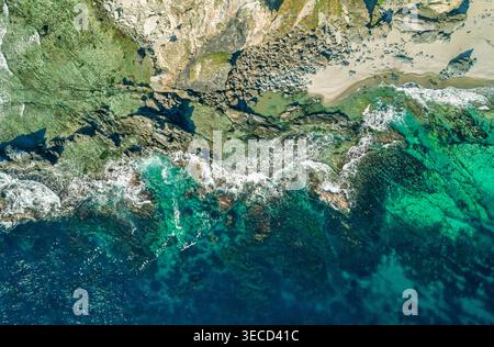 Aus der Vogelperspektive fängt leuchtende blaue und grüne Meereswellen auf eine felsige Küste und weißen Sandstrand an einem sonnigen Tag ein Stockfoto