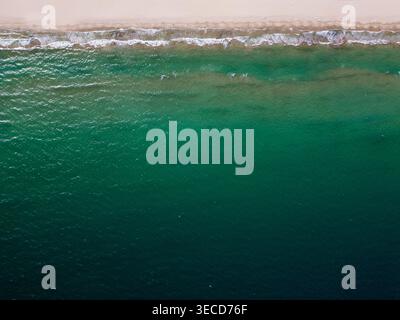 Hochwinkel-Drohnenaufnahme einer wunderschönen Sandküste, die auf das tiefblaue Meer trifft. Die Top-down-Perspektive zeigt kleine weiße Wellen, die auf dem abstürzen Stockfoto