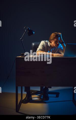 Mann, der spät am Laptop arbeitet, sieht erschöpft und überfordert aus im dunklen Büro. Stockfoto