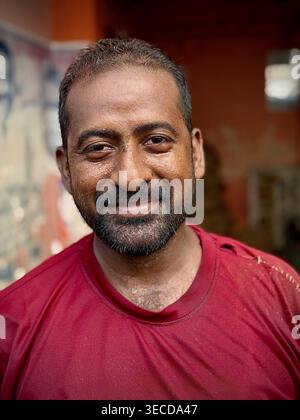Ein freundlich lächelnder Mann, mit Schweißperlen im Gesicht, in einem roten Pullover mit Bart vor verschwommenem Hintergrund, Indien Stockfoto