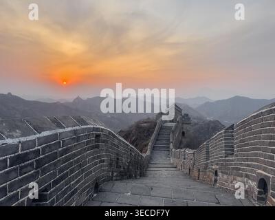 Der Sonnenuntergang leuchtet über alten Steinmauern. Treppen führen nach oben durch zerklüftete Berggipfel. Goldenes Licht färbt den Himmel in sanften bernsteinfarbenen Tönen. Weit entfernte Türme stehen stolz inmitten nebeliger Hügel Stockfoto