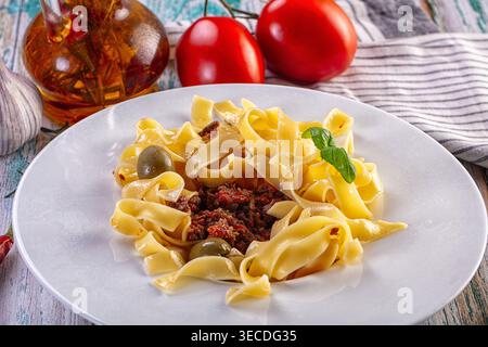 Appetitliche italienische Tagliatelle mit Fleischsoße, Oliven und Basilikum. Frische Tomaten und Knoblauch im Hintergrund auf einem rustikalen Tisch. Ein warmes, inv Stockfoto