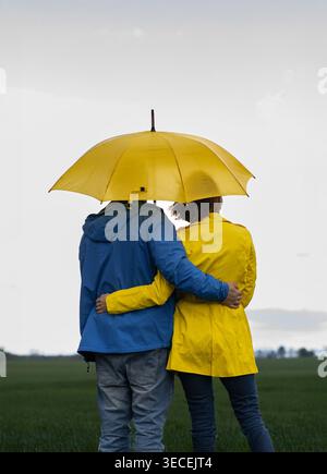 Rückansicht eines Paares, Mann in blauer Jacke, Frau in gelbem Regenmantel, umarmt unter einem großen, hellgelben Regenschirm draußen. Vorstellung von Liebe, gegenseitigem Beistand Stockfoto