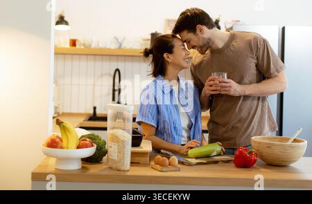 In einer hellen Küche teilen sich eine Koreanerin und ein Kaukasier einen Moment beim Kochen. Sie stehen neben einem Schneidebrett mit Gemüse und genießen jedes o Stockfoto