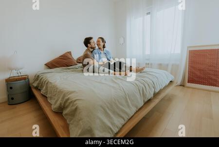 Auf einem großen Bett sitzen eine Koreanerin und ein kaukasischer Mann in einem sonnigen, luftigen Zimmer zusammen. Stockfoto