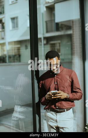 Ein Mann steht neben einer Glaswand in einer Stadt. Er schaut mit einem Lächeln auf sein Handy. Die Reflexionen des Fensters zeigen Teile des Gebäudes und der Straße Stockfoto