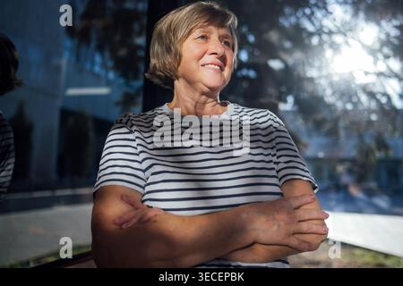 Eine ältere Frau mit kurzen Haaren in einem gestreiften Hemd sitzt mit überkreuzten Armen und lächelt in der Nähe eines großen Fensters mit Sonnenlicht, das die Szene beleuchtet Stockfoto