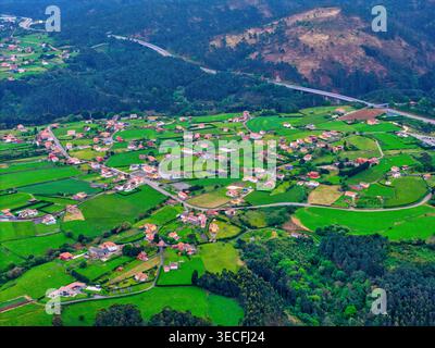 Blick aus der Vogelperspektive auf ein ländliches Dorf mit Häusern, die über lebendige grüne Felder und bewaldete Hügel in der Nähe eines Autobahnviadukts in Cudillero, Asturias, Spa verstreut sind Stockfoto
