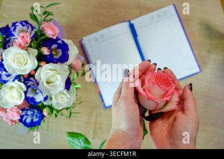 Weibliche Hände halten eine Rosenblume. Notizbuch, Tagebuch mit einem Stift auf einem Holztisch in der Nähe eines Straußes aus mehrfarbigen Rosen im Hintergrund. Draufsicht, flach Stockfoto