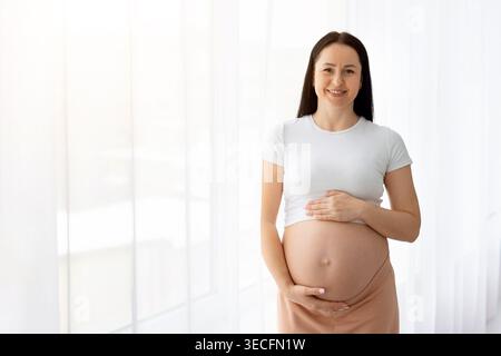 Schwangere junge kaukasische Frau lächelt, während sie den Bauch hält und vor hellem Hintergrund steht Stockfoto