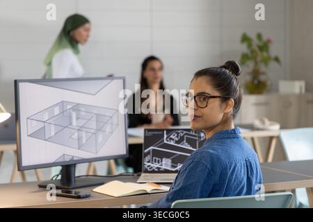 Asiatische Frau in Denim-Hemd, die sich am offenen Schreibtisch zur Kamera dreht und den 3D-Grundriss auf dem Monitor überprüft. Büro, Zusammenarbeit, Architektur, Design, Miete Stockfoto
