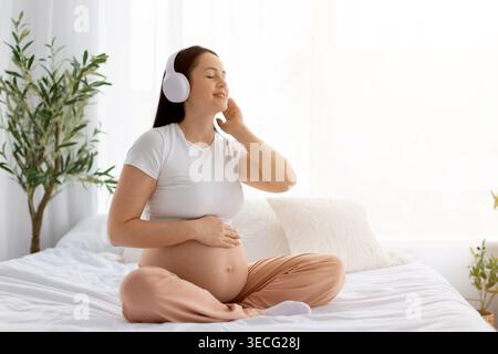 Schwangere Frau, die auf dem Bett sitzt und Musik in weißen kabellosen Kopfhörern hört Stockfoto