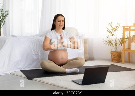 Schwangere junge Frau, die zu Hause Yoga übt, mit Laptop davor Stockfoto