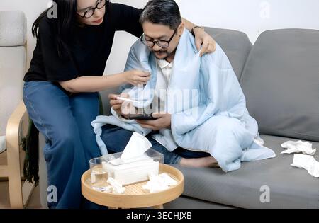 Diese Indoor-Szene veranschaulicht einen mitfühlenden häuslichen Moment, in dem eine Frau einem Mann hilft, der an einer Erkältung oder Grippe leidet, während er seine t überwacht Stockfoto