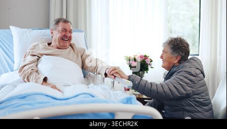 Lachen, altes Paar und Hände im Krankenhaus halten für Unterstützung, lustige Unterhaltung und Besuch für Komfort. Lächeln, Sorgfalt und Senioren mit Humor Stockfoto