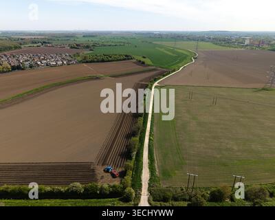 Aus der Vogelperspektive sehen Sie riesige landwirtschaftliche Felder mit einem Traktor pflügenden Boden, einen geraden ländlichen Weg und Wohnhäuser unter klarem Himmel in der Nähe von Ferrybridge Stockfoto