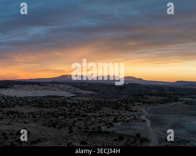 Aus der Vogelperspektive auf eine gewundene, unbefestigte Straße durch eine riesige Wüstenlandschaft mit schneebedeckten Bergen unter einem lebendigen Sonnenuntergangshimmel. Stockfoto