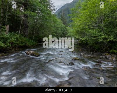 Aus der Vogelperspektive auf einen schnell fließenden Fluss mit Wildwasserschnellen, die sich durch einen üppigen grünen Wald und ein Bergtal mit einer kleinen Holzbrücke unter einem schlängeln Stockfoto