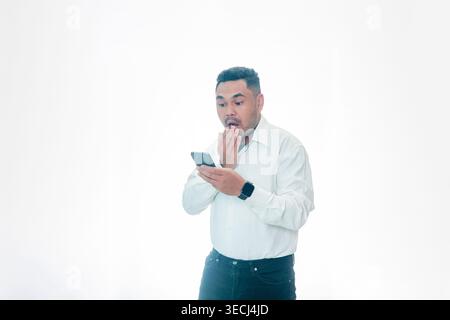Ein schockierter indonesischer Mann in einem weißen Hemd, der sich den Mund zuhält, während er auf sein Handy schaut, und Unglauben oder Überraschung ausdrückt, isoliert auf Weiß. Stockfoto