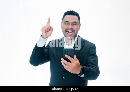 Ein glücklicher asiatischer Geschäftsmann in einem schwarzen Anzug, der ein Telefon hält und mit inspiriertem Ausdruck nach oben zeigt, isoliert auf einem sauberen weißen Hintergrund. Stockfoto