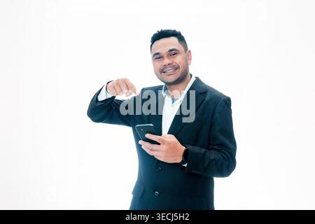 Ein glücklicher indonesischer Mann in einem schwarzen Anzug hält ein Smartphone und zeigt mit dem Finger auf das Gerät, das eine Empfehlung auf weißem Hintergrund anzeigt. Stockfoto