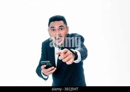 Ein indonesischer Mann in einem schwarzen Anzug blickt mit schockiertem Ausdruck in die Kamera, während er ein Smartphone hält und mit dem Finger nach vorne zeigt. Stockfoto