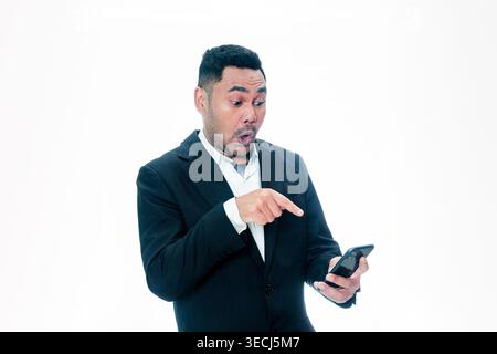 Ein indonesischer Mann in einem schwarzen Anzug, der mit schockiertem Gesichtsausdruck auf sein Handy schaut und mit dem Finger auf den Bildschirm zeigt, vor weißem Hintergrund. Stockfoto