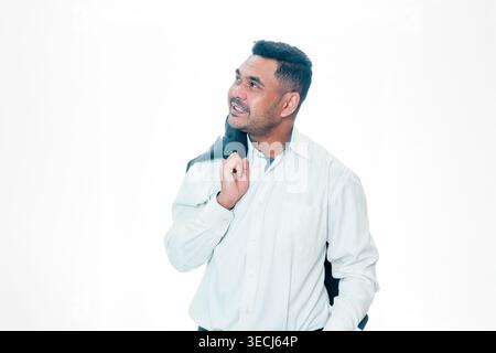 Glücklicher indonesischer Mann in einem weißen Hemd, der nachdenklich nach oben schaut, mit einem Anzugjackett über der Schulter, isoliert auf weißem Hintergrund. Stockfoto