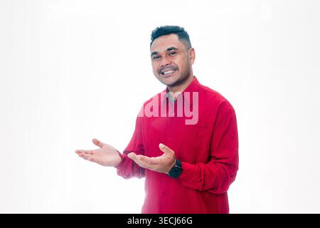 Lächelnder indonesischer Mann in einem roten Hemd, der mit offenen Handflächen etwas zur Seite zeigt auf weißem Hintergrund, perfekt für Kopierplatz. Stockfoto