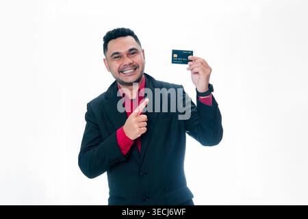 Porträt eines glücklichen indonesischen Mannes in einem Anzug, der eine Kreditkarte hält und darauf zeigt, isoliert auf weißem Hintergrund. Stockfoto