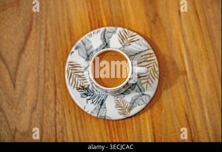 Traditioneller türkischer Kaffee, serviert in einer eleganten Porzellantasse auf einem Holz-Hintergrund, begleitet von einem kleinen Glas Likör oder Wasser. Authentischer Kaff Stockfoto