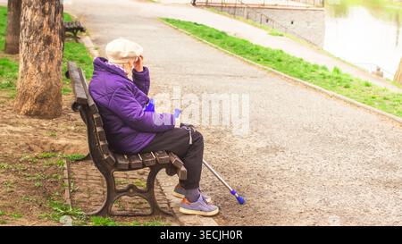 Eine ältere Frau in einer lila Jacke, die auf einer Holzbank mit blauen Ellenbogenkrücken im Freien liegt und nach unten blickt, während sie ihren Kopf in nachdenklicher Stimmung hält. Stockfoto