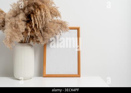 Vertikaler Holzrahmen mit weißer Vase mit trockenen Blumen über weißer Wand. Modellvorlage für Ihr Design, Text. Stockfoto