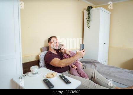 Glücklicher Vater und Tochter, die sich im Bett entspannen und ein Selfie mit einem Smartphone machen Stockfoto
