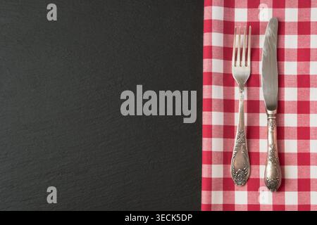 Besteck mit rotem kariertem Handtuch und leerem Etikett auf schwarzem Schieferhintergrund. Draufsicht. Menükarte für Restaurants. Stockfoto