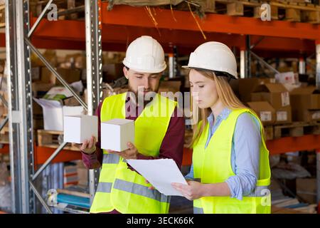 Männlicher Mitarbeiter zeigt ein weißes Kästchen, während weibliche Kollegin die Dokumentation eingeht. Einführung neuer Produkte und Bestandsaufnahme im Lagerbetrieb Stockfoto