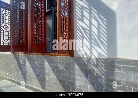 Das traditionelle Holzfenster wirft scharfe Schatten auf die graue Ziegelwand. Sonnenlicht tanzt durch komplizierte Gittermuster. Die Architektur verbindet Handwerkskunst mit natürlichem Licht. Ideal für historische Szenen. Kopierbereich. Stockfoto