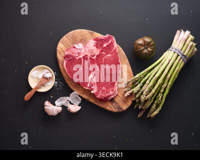 Auf einem Holzschneidebrett sitzt ein roh geschnittenes T-Bone-Steak neben frischem Spargel und einer Tomate. An der Seite befindet sich eine kleine Schüssel mit Salz und Knoblauch. Stockfoto