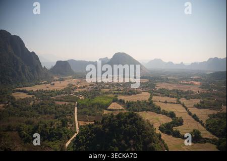 Panoramablick auf Vang Vieng, Laos, mit einem weiten Tal aus Feldern und Wäldern, umgeben von Kalksteinkarstbergen unter einem blassblauen Himmel und sanften Morgen Stockfoto
