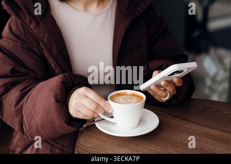 Nahaufnahme einer Person in einer warmen Jacke, die einen Cappuccino hält und ein Smartphone an einem Café-Tisch verwendet, um eine gemütliche Kaffeepause, einen Lifestyle und einen Moder festzuhalten Stockfoto