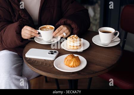 Nahaufnahme eines gemütlichen Kaffeetisches mit zwei Tassen Kaffee, süßem Gebäck und Tarte auf weißen Tellern und einem Smartphone in einem Etui für eine entspannte Winterkulisse Stockfoto