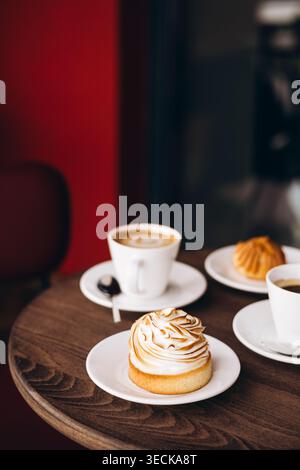 Gemütliche Café-Szene mit einer Tasse Kaffee auf einem Holztisch, serviert mit einer gerösteten Meringue-Tarte und einem kleinen Gebäck. Warme Farben, geringe Schärfentiefe, r Stockfoto